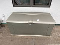 Front view of Rubbermaid outdoor storage box against a wall on a concrete patio surface showing minor dirt marks on lid and front.