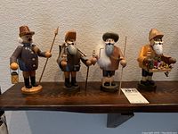 Four German wooden smoker figurines displayed on wooden shelf, each holding different props such as lantern, spear, clay pots, staff, and tray.