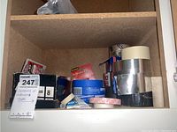Shelf with multiple rolls of duct tape (silver, black, white), painter's masking tape, 3M blue tape, red and white adhesive tape, and box of JB Weld epoxy adhesive.