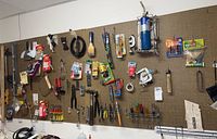 Wide view of assorted tools hanging on brown pegboard, showing hand saws, screwdrivers, pliers, torch, and hardware items.
