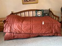 Full view of the twin sized wooden daybed with a rust-colored comforter and three decorative pillows, showing the bed frame design and protective brass finials.