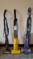 Three upright vacuum cleaners lined up: Bissell 3-in-1 on left, yellow Eureka The Boss center, purple Shark on right. Power cords wrapped on handles.