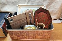 Wooden basket, plastic serving tray, serving board, decorative plate, coasters and coffee mug arranged inside the basket, showing overall lot contents.