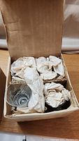 Cardboard box containing eight clear crystal glasses, each individually wrapped with paper padding inside compartmentalized sections.