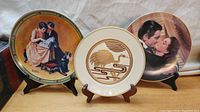 Three decorative plates on display stands: golden heron design on white background, Norman Rockwell style illustration of couple with dog, and vintage romantic couple plate.