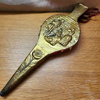 Front view of the brass plated fireplace bellows with embossed decorative figures and floral details on nozzle.