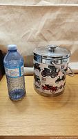 Ice bucket shown next to a water bottle for scale, displaying vintage car graphics and stainless steel handle.