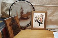 Lot with three Indigenous art pieces: wooden moose on leather background in wooden stick frame, wooden carved bird panel, framed Indigenous whale print