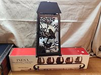 Metal lantern candleholder with bird and branch cutout design, placed on its box showing size and branding information