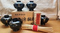 5 black lacquer Japanese rice bowls with gold design and lids arranged around a wooden box with Japanese script. Bundle of chopsticks in front of the bowls.