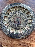 Front view of round metal plate with detailed peacock and floral motifs with segmented border design in gold, black, and red colors