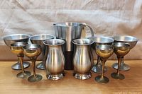 Front view of the lot showing all 11 pieces: one pitcher, two tumblers, four pewter goblets, and four brass cups arranged in three rows.