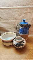 Photo showing all three pottery items on a wooden surface: a black and white vase with abstract design; a large blue and brown jug; and a white ceramic dish with colored bands.