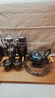 Lot of silver plate items on wooden surface including tall pitcher, jar, two small pots, covered dish and ornate tray with handles