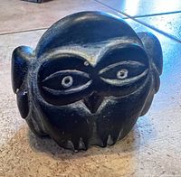 Photo of a small hand-carved owl figurine made of dark stone with white carved detailing on eyes, nose, and beak. Figurine shows minor nicks and wear.