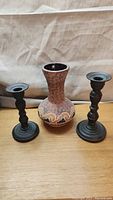 Pottery vase with crackle pattern and beige waves centered between two dark metal candle sticks with turned design on a wooden surface.