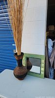 Photo showing two tone seagrass vase filled with tall natural stalks next to a small wooden framed mirror leaning against a wall.