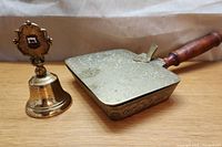 Vintage brass silent butler crumb catcher with wooden handle and floral embossed brass tray next to a vintage brass bell with ornate top on a neutral background.