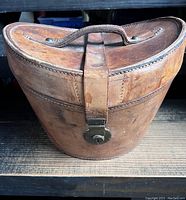 Closed view of the vintage leather hat box showing exterior wear, leather handle on top, and metal lock front.