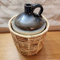 Front angled view of Western stoneware jug inside woven wicker basket cover on wooden surface.
