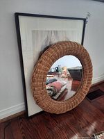 Round wicker framed mirror leaning on wall on top of a framed abstract art print, shows natural woven texture and reflective glass.
