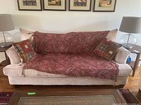 Front view of the full couch showing beige faux suede upholstery, two decorative cushions on each side, and a paisley patterned throw covering the seating area.