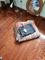 Covered folded coverlet with black embroidered table cover on top, placed on wooden floor next to lamps and round mirror.