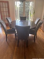 Six grey faux suede dining chairs arranged around a circular wooden dining table in a sunlit room with hardwood floors.