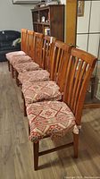 Row of six wooden dining chairs with slatted backrests and patterned cloth cushions, placed on a hardwood floor in a living room setting.