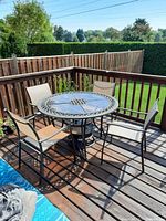 Four metal framed exterior chairs with beige mesh fabric seating placed around a round patio table on a wooden deck. Green lawn and fenced backyard in background.