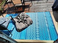 A blue patterned outdoor rug with tribal and geometric designs placed on a wooden deck. Two photos with slightly different angles show the full rug and some patio furniture and cushions partially on the rug.
