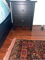 Front view of black IKEA night table positioned beside bed, showing two drawers with round knobs, wood floor and part of rug visible