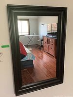 Composite framed wall mirror hanging on wall with reflection showing part of a room including furniture and window.