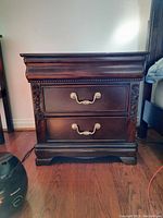 Front view of dark wood night table showing two drawers with metal handles and carved floral decoration on sides.