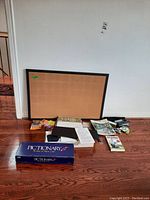 Photo showing framed cork board leaning on wall with Pictionary game box, books, papers, and various small office supplies spread on floor.