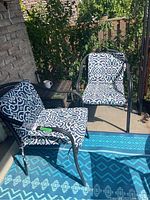 Two matching metal frame outdoor chairs with resin seats and backs, each outfitted with cushioned covers in a white, blue, and green geometric pattern. Chairs positioned outdoors by a brick wall and patio.