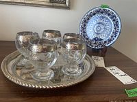 Overall view of the five glasses arranged on the silver plate tray with the decorative plate on wooden stand behind them.