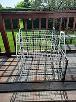 Front view of two white metal shoe racks arranged side by side on a wooden deck, showing the wireframe design and three shelves each.