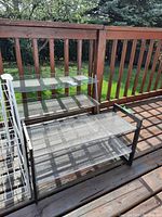 Two metal shoe racks placed outside on a wooden deck with a green lawn and wooden railing in background. The racks have mesh metal shelving and black frame.