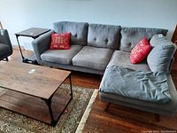 Side view of gray upholstered L-shaped modular couch with tufted cushions and red decorative pillows, sitting on a hardwood floor beside a wooden coffee table and a side table with wheels.