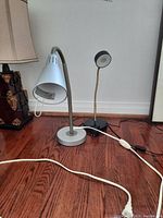Two task lamps side by side on wood floor showing one with flexible neck and white shade, the other smaller with rigid neck and round shade, plus cords and switches.