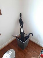 Three black umbrellas placed inside a metal umbrella holder positioned in a corner on a hardwood floor.