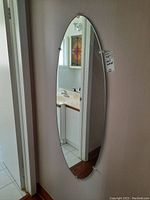 Front left angled view of an oval frameless wall mirror mounted vertically on the wall.