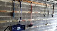 Photo of five different wall mounted tools, showing them clearly hung on wooden slats attached to a garage brick wall.