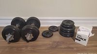 Two adjustable dumbbells with black weight plates and threaded collars, several loose weight plates stacked and separated, and a box of lifting chalk on the floor against a light wall.