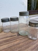 Four clear glass Mason jars with metal lids, two smaller and two larger. All show embossed lettering.