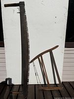 Photo showing both antique saws propped against a wall in daylight: a large crosscut saw with big teeth and a wooden handle, and a wooden framed bow saw with tension rods.