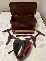 Open antique wooden sewing chest with four detached wooden legs, four large wooden clamps, a heart-shaped wooden container, and five vintage wooden thread spools in different colors placed on a mattress.