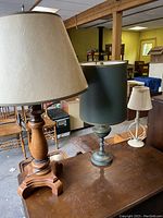 Three lamps on wood surface: wooden lamp with beige fabric shade, metal lamp with black cylindrical shade, small white lamp with round base and small cream shade