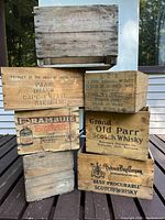 Front and side view of stacked assorted wooden liquor crates showing brand logos and text for Grand Old Parr Scotch Whisky, Drambuie, Hudson's Bay Company, and others.